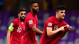 L'image montre des joueurs de football célébrant un moment de victoire ou d'accomplissement sur le terrain. Ils portent des maillots rouges avec des logos de l'équipe, et l'un d'eux semble montrer un symbole ou une médaille en soulevant son maillot. Les autres joueurs réagissent joyeusement derrière lui, montrant l'esprit d'équipe et la camaraderie. L'arrière-plan fait référence à un stade, créant une atmosphère d'événement sportif.