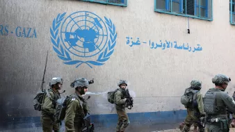 L'immagine mostra un gruppo di soldati in uniforme militare che camminano davanti a un edificio con un logo delle Nazioni Unite, situato nella regione di Gaza. I soldati indossano elmetti e sono equipaggiati per situazioni di operazioni di sicurezza. L'atmosfera è seria e il contesto suggerisce una situazione di tensione o di operazioni militari nella zona. Sul muro dell'edificio si vedono scritte in arabo e in inglese, che indicano che si tratta di un ufficio dell'UNRWA, l'Agenzia delle Nazioni Unite per i Rifugiati Palestinesi.