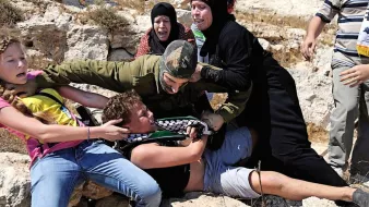 L'image montre une scène de tension où plusieurs personnes semblent être impliquées dans une confrontation. Un militaire en uniforme est en train de maîtriser un jeune homme au sol tandis que d'autres personnes, dont des femmes et des enfants, semblent réagir avec de l'indignation ou de la peur. L'environnement paraît rural, avec des rochers et une végétation sèche. L'émotion et le conflit sont palpables dans cette image.