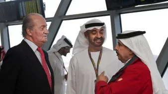 The image shows a gathering of three men engaged in conversation. Two of them are dressed in traditional attire, typical of Middle Eastern cultures, with one wearing a white kandura and the other in a black and white headdress. The third man is in formal Western attire with a suit and a red tie. The background suggests an airport or a similar setting, indicated by the large windows and digital displays. The expressions of the men appear to be friendly and engaged in a dialogue.