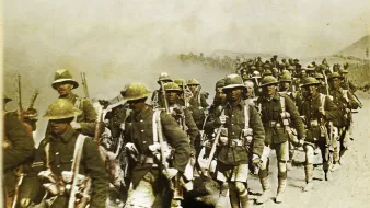 L'image représente un groupe de soldats en marche, probablement pendant une période de conflit. Ils portent des uniformes militaires, y compris des casques et des équipements de combat. Les soldats avancent sur un terrain poussiéreux, et il semble qu'ils soient dans un environnement difficile, avec une atmosphère chargée de fumée ou de poussière. L'image donne une impression de détermination et de camaraderie parmi les troupes.