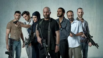 L'image montre un groupe de sept personnes armées, se tenant debout sur un fond gris. Ils ont des expressions déterminées et semblent en position de confrontation. Certains d'entre eux tiennent des armes à feu, tandis qu'une femme porte un hijab et se tient au milieu du groupe. Les hommes sont vêtus de vêtements variés, allant de casual à militaire. L'atmosphère est intense et pourrait suggérer un contexte d'action ou de tension.