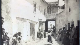 L'image représente une scène de rue dans un quartier de Constantine, en Algérie. On y voit des maisons blanches avec des volets, et une ruelle pavée qui semble étroite. Plusieurs personnes, y compris des enfants, sont visibles, certaines marchant et d'autres se tenant debout. Les vêtements des habitants semblent traditionnels, reflet d'une époque passée. L'atmosphère générale évoque une vie quotidienne animée dans un contexte historique.
