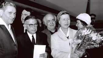 The image captures a group of individuals outside an airplane, likely at an airport. There are six people in the frame, with one woman prominently holding a bouquet of flowers. She appears to be the center of attention, dressed in a light-colored outfit and wearing a headscarf. The men around her are dressed in suits and vary in expressions. The background includes the tail of the aircraft, suggesting a public or ceremonial occasion. The image is in black and white, giving it a classic, historical feel.