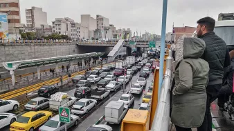 تصویر نشان‌دهنده یک صحنه شلوغ از ترافیک در یک خیابان شهری است. در سمت چپ تصویر، افرادی در حال تماشا از بالا ایستاده‌اند. در پایین، خطوط خودروها به وضوح قابل مشاهده است که نشان‌دهنده تجمع و ترافیک سنگین است. بقیه عناصر شهری مانند ساختمان‌ها و علائم راهنمایی نیز در پس‌زمینه دیده می‌شوند. هوای ابری و احتمالا بارانی به دید فضا و حس و حال تصویر اضافه می‌کند.