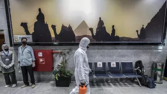 تظهر في الصورة مشهداً داخل محطة أو مطار. في الخلفية يوجد جدار مزين بصورة لأهرامات مصر مع silhouettes لحيوانات وحشود. في المقدمة، يسير شخص يرتدي بدلة واقية وقناع، حاملاً حقيبة برتقالية. بجانبه، يوجد موظفان يرتديان كمامات ويتوليان خدمات الاستقبال. في الزاوية، توجد مقاعد فائضة وبعض النباتات. تعكس الصورة أجواء احترازية مع لمسة ثقافية.