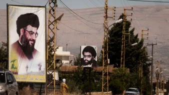 L'image montre une rue avec des panneaux d'affichage sur lesquels figurent des portraits d'un homme portant des lunettes et une barbe, associé à une ambiance rurale. On peut également voir des lignes électriques et quelques bâtiments en arrière-plan. À gauche, un panneau est en couleur et à droite, un autre est en noir et blanc. La scène semble être située dans une région montagneuse. C'est une illustration d'un espace public où des affiches politiques ou religieuses peuvent être observées.