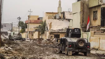 الصورة تظهر مشهداً من منطقة متضررة بسبب نزاع أو حرب. هناك الكثير من الخراب في المباني المحيطة، حيث تبدو العديد منها مدمرة أو شبه مدمرة. في منتصف الصورة، توجد مركبة عسكرية، يبدو أنها تُستخدم في عمليات الأمن أو الاستجابة الطارئة. تلوح في الأفق مئذنة مسجد، مما يشير إلى وجود مجتمع محلي. الشوارع تبدو مليئة بالتراب والحطام، مما يعكس آثار الدمار الذي لحق بالمنطقة.