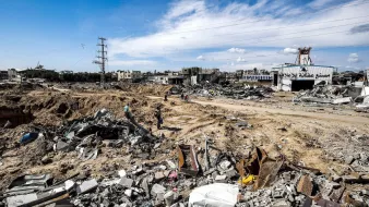 L'immagine mostra un paesaggio devastato, caratterizzato da macerie e detriti. Il terreno sembra essere stato scavato e distrutto, con edifici in rovina visibili sullo sfondo. I resti di strutture crollate sono sparsi ovunque, mentre alcune persone si muovono tra le macerie. Il cielo è parzialmente nuvoloso, e ci sono segnali di infrastrutture elettriche nei dintorni. L'atmosfera trasmette un senso di desolazione e distruzione.