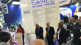 تظهر الصورة مجموعة من الأشخاص في حدث أو معرِض، حيث يتواجد متحدثة أمام منصة تحمل شعار "إسرائيل" وتفاصيل عن الجناح الوطني. هناك بعض الحضور في الخلفية يستمعون بانتباه، بينما يبدو بعضهم منشغلاً بتدوين الملاحظات. المنصة محاطة بالزهور، ويوجد مرافقون يرتدون بدلات رسمية. الأجواء تعكس نشاطًا رسميًا ومهنيًا.