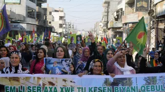 تصویر یک تظاهرات بزرگ را نشان می‌دهد که گروهی از زنان در آن شرکت دارند. آنها در خیابانی شلوغ راه می‌روند و پلاکاردهایی به دست دارند. تعدادی پرچم با نمادهایی خاص نیز در دست دارند. زنان با لبخند و دست‌های بالا به نشانه حمایت و ابراز احساسات مشارکت می‌کنند. محیط اطراف شامل ساختمان‌ها و جمعیت دیگری است که در پس‌زمینه دیده می‌شود. این تصویر به وضوح نشان‌دهنده یک حرکت اجتماعی یا سیاسی است.