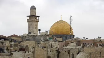 L'image montre une vue d'un paysage urbain avec des bâtiments en pierre. Au centre, on peut apercevoir un dôme doré, probablement celui du Rocher, qui est un site religieux important. À proximité, on remarque un minaret qui surplombe les structures anciennes. Le ciel est nuageux, ajoutant une atmosphère douce à la scène. Les toits sont parsemés de paraboles satellites, témoignant de la modernité mêlée à l'histoire de cette région.
