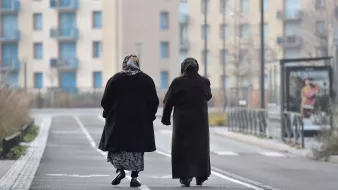 L'image montre deux femmes marchant côte à côte sur une voie urbaine. Elles portent des manteaux sombres et des foulards, ce qui suggère qu'il fait frais. En arrière-plan, on distingue des bâtiments modernes avec des balcons et des fenêtres. La scène dégage une atmosphère tranquille, avec un chemin bordé d'herbes. Leurs silhouettes semblent illustrer un moment de complicité et de camaraderie.