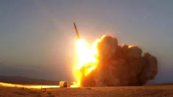 L'image montre le lancement d'un missile. On peut voir une grande flamme et une traînée de fumée qui s'échappent du sol alors que le missile s'élève dans le ciel. L'environnement semble désertique, avec un éclairage crépusculaire qui souligne la force de l'explosion et le mouvement ascendant du projectile.