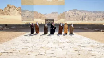 تظهر في الصورة مجموعة من الأشخاص يرتدون الزي التقليدي، ويقفوا على طريق مرصوف بالحجارة في منطقة صحراوية. خلفهم توجد جدران عاكسة تعكس المنظر الجبلي المحيط، مما يعطي شعوراً بالتواصل بين البشر والطبيعة. السماء زرقاء وصافية، مما يضيف جمالاً على المشهد.