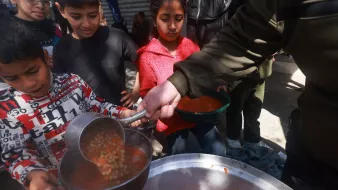 در این تصویر، گروهی از کودکان در حال دریافت غذا هستند. یکی از کودکان که لباس‌های رنگارنگ دارد، با دقت در حال ریختن خورش یا سوپ به ظرفش است. در پس‌زمینه، سایر کودکان نیز منتظر هستند تا نوبتشان برسد. محیط بیرونی به نظر شلوغ و غیررسمی است و نشان‌دهنده یک فعالیت جمعی برای سرو غذا به افراد نیازمند می‌باشد.