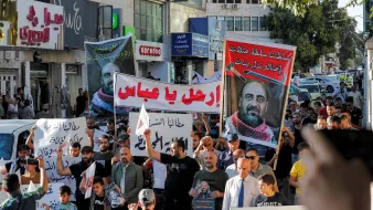 L'image montre une foule de manifestants dans une rue. Les participants portent des pancartes et des banderoles avec des écrits en arabe. L'ambiance semble être celle d'une manifestation ou d'un rassemblement public. Plusieurs personnes brandissent des affiches, et on peut voir des voitures et des bâtiments en arrière-plan. Les manifestants semblent engagés et déterminés dans leur action.