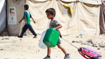 L'immagine mostra un bambino che cammina in un ambiente simile a un campo profughi. Indossa una maglietta a righe e pantaloni corti. Porta con sé un contenitore verde e una valigia rosa con ruote. Sullo sfondo, si possono vedere altre persone e tende di un campo, il che suggerisce una situazione di emergenza. Il terreno sembra polveroso e desolato. Il bambino appare concentrato mentre si sposta.