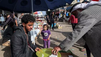 در تصویر، یک بازار محلی به چشم می‌خورد. در وسط تصویر، مرد جوانی نشسته و در حال فروش کالاهایی است. او یک بشقاب سبز رنگ را در دست دارد که در آن چند قلم وجود دارد. به نظر می‌رسد که او به مشتری‌اش توجه دارد که در حال انتخاب کالاهاست. در کنار آن، یک دختر بچه نیز در حال بازی کرده و به صحنه نگاه می‌کند. در پس‌زمینه، افراد دیگری در حال خرید و گشت و گذار هستند و جوی شلوغ و پرجنب‌وجوش به چشم می‌خورد. سایه چترهایی که بالای سر برخی افراد است، فضایی دلپذیر ایجاد کرده است.