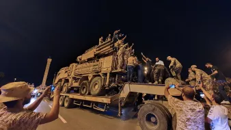 L'immagine mostra un gruppo di persone radunate intorno a un veicolo militare, che sembra essere un camion con un modulo di artiglieria. La scena si svolge di notte, con luci artificiali che illuminano l'ambiente. Alcuni individui sono saliti sopra il veicolo, mentre altri stanno scattando foto con i loro telefoni. L'atmosfera sembra carica di emozione e partecipazione, con gesti di saluto o di celebrazione da parte delle persone sopra il veicolo.