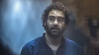 L'immagine mostra un uomo con capelli ricci e occhiali. Indossa una maglietta o camicia scura, ed appare sorpreso o preoccupato. Lo sfondo è sfocato e contribuisce a un'atmosfera di tensione o intensità. L'illuminazione è tenue, dando un effetto drammatico alla scena.