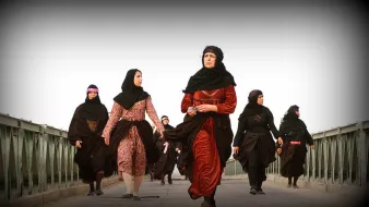 L'image montre un groupe de femmes marchant sur un pont. Elles portent des vêtements traditionnels, principalement des robes longues et des foulards. La scène semble être en extérieur, avec un ciel clair en arrière-plan. Les femmes ont des expressions déterminées et se déplacent ensemble, ce qui dégage une impression d'unité et de force.