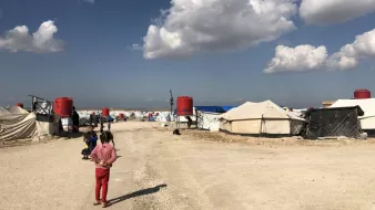 تظهر في الصورة مخيم للاجئين أو النازحين، حيث تتواجد عدة خيام مرتبة على جانبي الطريق. السماء صافية مع وجود سحب بيضاء، والأرض مرصوفة بالحجارة. يمكن رؤية بعض الأطفال يلعبون في المنطقة. يبدو أن المخيم يعاني من ظروف بسيطة، مما يشير إلى حياة موقتة للأشخاص المقيمين فيه.