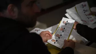 The image shows a person, partially visible, holding a ballot or voting card that features photographs and names of candidates. The setting appears to be a polling station, with multiple ballots or documents visible. The individual seems to be examining the options before making a decision. The lighting suggests an indoor environment, likely during a voting process.