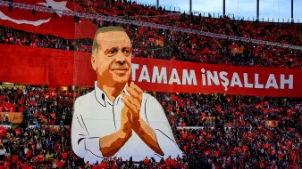 L'image montre une grande scène dans un stade rempli de supporters. Au centre, il y a une immense illustration d'un homme qui semble être un leader ou un personnage politique, probablement Erdoğan, qui applaudit. En arrière-plan, une banderole rouge avec des inscriptions en lettres blanches affiche "TAMAM İNŞALLAH". Les spectateurs tiennent des drapeaux rouges, créant une atmosphère colorée et festive. L'ensemble évoque une manifestation de soutien, probablement lors d'un événement politique.