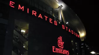 L'image montre l'extérieur de l'Emirates Stadium, qui est la maison du club de football Arsenal, à Londres. Le bâtiment est illuminé, avec le nom "EMIRATES STADIUM" en lettres rouges lumineuses bien visibles. On peut également voir le logo d'Emirates, une compagnie aérienne, mis en évidence. L'ambiance semble animée, suggérant qu'il y a probablement un événement ou un match en cours, avec des personnes rassemblées autour de la façade du stade.