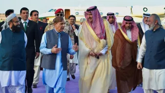 L'image montre un groupe de personnes en tenue formelle, marchant ensemble sur un tapis violet. On peut voir des hommes portant des costumes traditionnels et des vêtements d'apparat, avec certains en toges ou en robes. En arrière-plan, un avion privé est stationné, indiquant un contexte officiel ou diplomatique. L'atmosphère semble conviviale, avec les participants engagés dans une conversation.