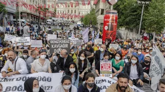 La imagen muestra una manifestación en una plaza urbana, con varias personas sentadas en el suelo. Los participantes sostienen pancartas y carteles que tienen mensajes de protesta. La mayoría de las personas llevan mascarillas, lo que puede indicar que la manifestación está ocurriendo en un contexto reciente de pandemia. Al fondo, se pueden ver banderas turcas y edificios, lo que sugiere que la protesta tiene lugar en Turquía. La atmósfera es de movilización y reclamo social.