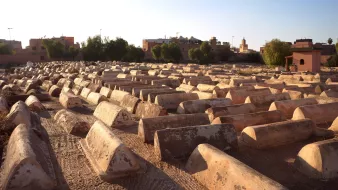 L'image montre un cimetière où l'on peut voir des tombes en forme de tombeaux, étalées sur un terrain sec. Les structures sont de couleur beige ou terre cuite et semblent être en béton, avec une surface usée par le temps. En arrière-plan, on aperçoit des bâtiments et des arbres, indiquant que le cimetière est situé près d'une ville. La lumière du soleil crée une atmosphère calme et paisible, tandis que le ciel est dégagé, laissant entrevoir un beau jour.
