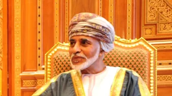 The image features a man seated in a richly decorated chair, wearing traditional attire. He is dressed in a white dishdasha, complemented by a decorative cape, and a turban, which is traditional in some Middle Eastern cultures. The background consists of intricate woodwork and ornate design elements, suggesting an opulent setting. The man's expression appears calm and thoughtful.