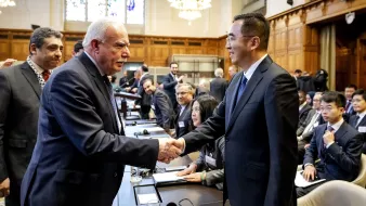 La imagen muestra a un grupo de personas en un entorno de reunión, probablemente en una conferencia o cumbre. En primer plano, dos hombres se están estrechando la mano, lo que sugiere un saludo cordial o un acuerdo. Uno de ellos tiene el cabello canoso y viste un traje oscuro, mientras que el otro es más joven y usa un traje también oscuro. Al fondo, se pueden ver otras personas sentadas alrededor de una mesa, con documentos y laptops, observando la interacción. El ambiente es formal y parece que se trata de un encuentro importante.