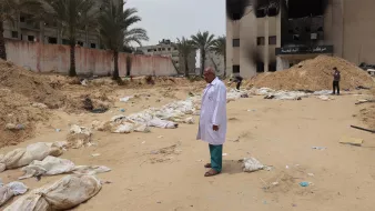 L'immagine mostra un paesaggio desolato e drammatico, caratterizzato da un terreno sabbioso. Al centro, un uomo in un camice bianco osserva un'area circondata da edifici danneggiati, con alcuni segni di distruzione visibili sullo sfondo. Intorno a lui ci sono sacchi bianchi disposti a terra, che suggeriscono una scena di grande sofferenza e perdita. Palme si ergono sullo sfondo, ma l'atmosfera generale è segnata da un senso di tristezza e desolazione.