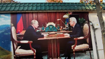 L'image montre deux hommes assis face à face à un bureau dans un bureau luxueux. Ils semblent s'engager dans une conversation sérieuse. Le décor est riche, avec des éléments de mobilier élégants et des accessoires de bureau. En arrière-plan, on peut apercevoir une grande fenêtre qui laisse entrevoir un paysage montagnard.