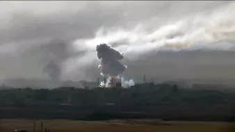La imagen muestra una escena con humo denso y nubes oscuras que se elevan desde un área urbana. Se aprecian instalaciones industriales o edificios en el fondo, y el humo parece provenir de una explosión o un incendio. El cielo está nublado, lo que contribuye a una atmósfera sombría y tensa en la imagen.