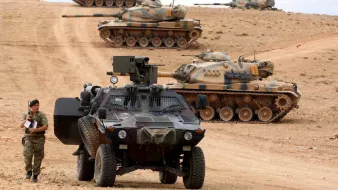 L'image montre un paysage militaire avec plusieurs chars d'assaut en arrière-plan, alignés sur une colline. Au premier plan, un véhicule blindé est garé sur un chemin poussiéreux, tandis qu'un soldat semble faire une inspection ou prendre des notes à côté du véhicule. Le terrain est désertique, et le ciel est partiellement nuageux, créant une atmosphère tendue et militaire. Les chars sont camouflés avec des motifs de peinture qui se fondent dans le paysage environnant.