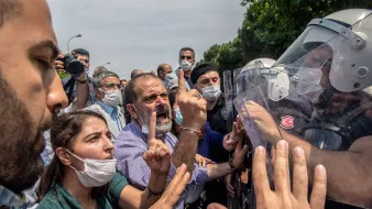 تظهر الصورة محتجين يتجمعون في مكان عام، حيث يبدو أن هناك توترًا في الأجواء. بعض الأشخاص يحملون لافتات، بينما يتواجد عدد من رجال الشرطة الذين يرتدون خوذات ودروع لمواجهة الحشد. يبدو أن هناك تفاعلًا حادًا بين المتظاهرين ورجال الأمن، مع تعبيرات وجهية تشير إلى الغضب والاحتجاج. بعض الأشخاص يرتدون أقنعة للوجه، مما يدل على احتمال وجود تدابير صحية. الأشجار والخضرة، تشير إلى أن الموقع في منطقة حضرية.