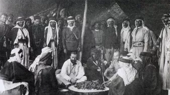 این تصویر تاریخی گروهی از مردان را در یک محیط سنتی نشان می‌دهد. آنها در یک چادر یا فضای باز گرد هم آمده‌اند و به نظر می‌رسد در حال تهیه یا صرف غذا هستند. چند نفر از آن‌ها لباس‌های سنتی عربی به تن دارند، از جمله عبا و headscarf (کشیه). در مرکز تصویر، چند نفر در حال نشستن و گفتگو هستند و سایرین ایستاده یا نشسته در اطراف آن‌ها قرار دارند. فضای تصویر گرم و صمیمی به نظر می‌رسد و نمایانگر فرهنگی غنی و سنت‌های اجتماعی است.