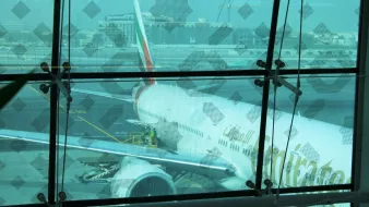 La imagen muestra un avión de la aerolínea Emirates estacionado en un aeropuerto. Se puede ver parte del fuselaje y el ala del avión a través de un gran ventanal con un diseño en las ventanas. Hay trabajadores en la plataforma que parecen estar realizando tareas de mantenimiento o preparación, y se puede observar un entorno aeroportuario con otros edificios y maquinaria en el fondo. La atmósfera es luminosa, aunque un poco difusa debido al cristal.
