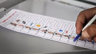 L'image montre une main tenant un stylo en train de remplir un bulletin de vote. Le bulletin affiche plusieurs colonnes, chacune contenant des photos de différentes personnes, accompagnées de symboles et de cases à cocher. Cela semble être un contexte électoral, où une personne vote pour des candidats. Le bulletin est posé sur une surface grise.