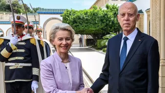 L'immagine mostra un incontro ufficiale tra due persone. Una donna, vestita con un abito chiaro, sorride mentre stringe la mano a un uomo in giacca e cravatta scura. Sullo sfondo, si possono vedere alcuni soldati in uniforme, che sembrano fare la guardia. La scena si svolge in un ambiente elegante, con piante e architetture raffinate visibili.