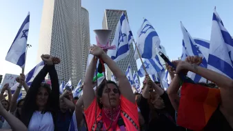 L'immagine mostra una folla di persone che partecipa a una manifestazione. Alcuni partecipanti tengono in alto delle bandiere israeliane, mentre molti altri alzano le mani, dimostrando un senso di unità e protesta. In primo piano, si può vedere una donna con una maglietta di colore neon che sembra guidare la manifestazione con un megafono. Sullo sfondo, si notano alti edifici moderni, suggerendo che l'evento si svolge in un contesto urbano. L'atmosfera è carica di energia e determinazione.