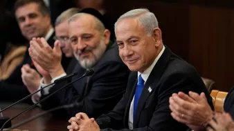 The image depicts a group of men seated around a table during a formal meeting. In the foreground, a man in a suit with a blue tie appears to be the focal point, wearing a slight smile. He is flanked by other men who are also dressed in formal attire. One man, likely a religious figure, is seen smiling broadly. The setting appears to be an official governmental or diplomatic environment, indicated by the wooden table and the serious demeanor of those present. There are papers and a microphone in front of them, suggesting they are engaged in a discussion or announcement.