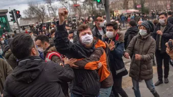 در تصویر، گروهی از جوانان در حال برگزاری یک تجمع هستند. برخی از افراد ماسک به صورت دارند و به نظر می‌رسد که در حال ابراز نظرات یا اعتراضات خود هستند. یکی از افراد به وضوح در حال فریاد زدن و دستش را به هوا بلند کرده است، در حالی که دیگران او را نگهداشته‌اند. به نظر می‌رسد وضعیت هیجانی است و برخی از افراد در تلاش برای برقراری نظم یا جلوگیری از درگیری هستند. پشت زمینه نیز شهری را نشان می‌دهد که با تابلوهای ترافیکی و ساختمان‌ها مشخص شده است.