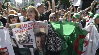 الصورة تظهر مجموعة من المتظاهرين في الجزائر، يحملون لافتات وأعلام. يبدو أن هناك نساء ورجال في المقدمة، ويعبرون عن مطالبات سياسية. إحدى اللافتات تحمل اسم "مصطفى عويصي" وعبارة "حرروا". الأجواء توحي بالحماسة والمطالبة بالتغيير.