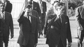 L'image montre un groupe de personnes marchant sur un chemin, avec plusieurs individus en costume. Deux hommes au premier plan saluent, vraisemblablement en se dirigeant vers une foule ou un événement. En arrière-plan, on peut apercevoir d'autres personnes et des éléments d'architecture, ainsi que des officiers en uniforme. L'atmosphère semble formelle et dynamique, indiquant une occasion spéciale.