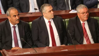 L'image montre trois hommes assis côte à côte dans un cadre formel, probablement lors d'une réunion ou d'une séance parlementaire. Ils portent des costumes sombres et des cravates, affichant une expression sérieuse. L'un d'eux porte une cravate rouge, tandis que les autres optent pour des nuances plus sombres. En arrière-plan, on aperçoit des personnes en costume, suggérant un environnement officiel.